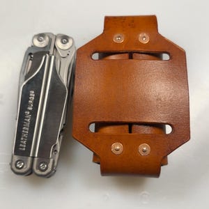 Könnte beinhalten: Ein silbernes Multi-Tool und eine braune Lederscheide. Auf dem Multi-Tool steht "LEATHERMAN SURGE". Die Scheide ist rechteckig mit zwei Schlitzen und Kupfernieten. Das Set liegt auf einer weißen Oberfläche.
