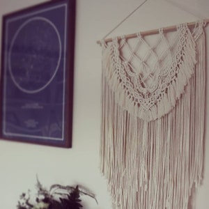 May include: A white macrame wall hanging with a geometric design and fringe. The hanging is made of cotton cord and is attached to a wooden dowel.
