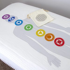 May include: A white massage table cover featuring a gray human figure outline with seven colorful chakra symbols. A small beige bag with a black geometric design rests on the cover. The symbols are purple, blue, green, yellow, orange, and red.
