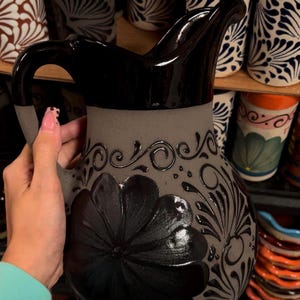 May include: A gray ceramic pitcher with a black floral design and handle. The pitcher has a glossy black top and a matte gray body with a large flower and swirling patterns. Other ceramic items are visible in the background.