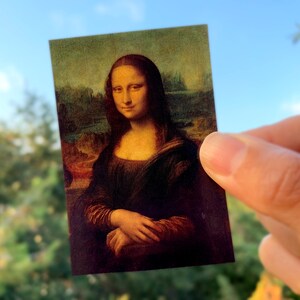 May include: A close-up of a reproduction of the Mona Lisa painting, a famous portrait by Leonardo da Vinci. The painting is on a card held by a hand.