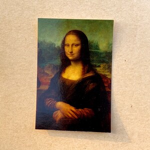 May include: A close-up of the Mona Lisa painting, a famous portrait of a woman with a mysterious smile. The painting is set against a landscape background with a rocky outcropping and a body of water.