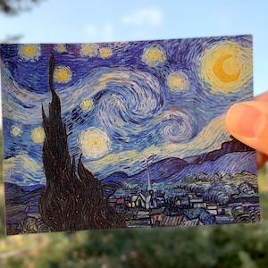 May include: A close-up of a postcard featuring a reproduction of Vincent van Gogh's famous painting, "The Starry Night". The painting depicts a swirling night sky with a bright yellow moon and stars, and a small village in the foreground.