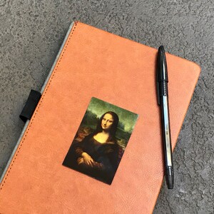 May include: A brown leather-bound journal with a black pen and a small image of the Mona Lisa on the cover.