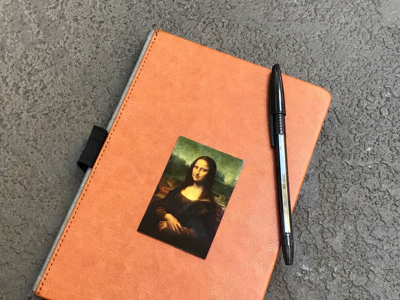 May include: A brown leather-bound journal with a black pen and a small image of the Mona Lisa on the cover.