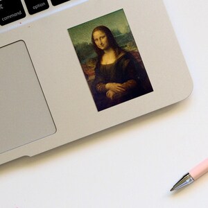 May include: A sticker featuring a painting of the Mona Lisa, a famous portrait by Leonardo da Vinci. The sticker is on a silver laptop.