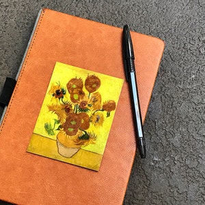 May include: A brown leather journal with a sunflower painting sticker on the front cover. A black pen is placed on the journal.
