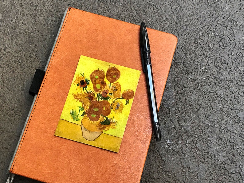May include: A brown leather journal with a sunflower painting sticker on the front cover. A black pen is placed on the journal.
