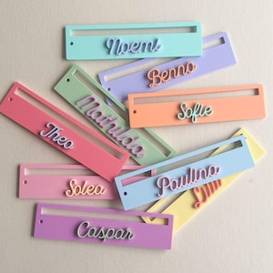 Könnte beinhalten: Eine Sammlung farbenfroher Namensschilder in verschiedenen Pastelltönen, darunter Rosa, Blau und Lila. Jedes Schild trägt einen anderen Namen in einer Schreibschrift. Die Schilder sind rechteckig mit einem Ausschnitt.