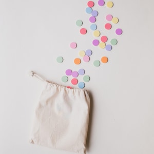 Könnte beinhalten: Eine weiße Baumwolltasche mit Kordelzug, aus der bunte runde Konfetti herausfallen. Das Konfetti ist in den Farben Rosa, Lila, Blau, Grün und Orange.
