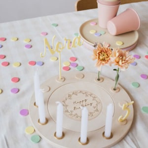 May include: A wooden birthday cake stand with a spinning top and candles. The stand has a white background with the words "Happy Birthday" engraved in the center. The stand is surrounded by colorful confetti.