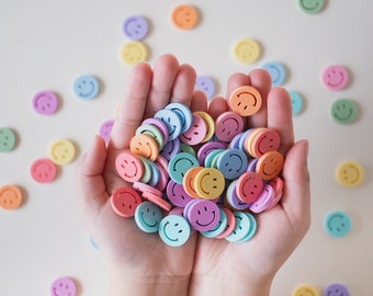 May include: A pair of hands holding a large pile of colorful smiley face charms. The charms are in various shades of pink, blue, yellow, and orange.