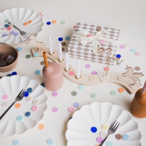 May include: A white table setting with two white plates, a brown bowl, a wooden unicorn, a wooden candle holder with white candles, a brown cup with a straw, and colorful confetti. The table is set for a birthday party.