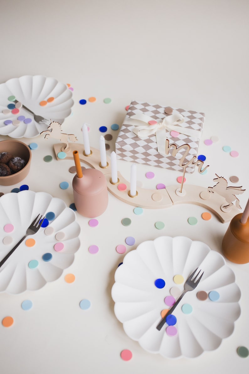 May include: A white table setting with two white plates, a brown bowl, a wooden unicorn, a wooden candle holder with white candles, a brown cup with a straw, and colorful confetti. The table is set for a birthday party.