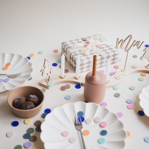May include: A birthday party table setting with white plates, a brown bowl with chocolate candies, a wooden candle holder with white candles, a gift wrapped in a brown and white checkered paper, a brown and orange silicone cup, and colorful confetti scattered on the table. The name "Mia" is written on a wooden sign.