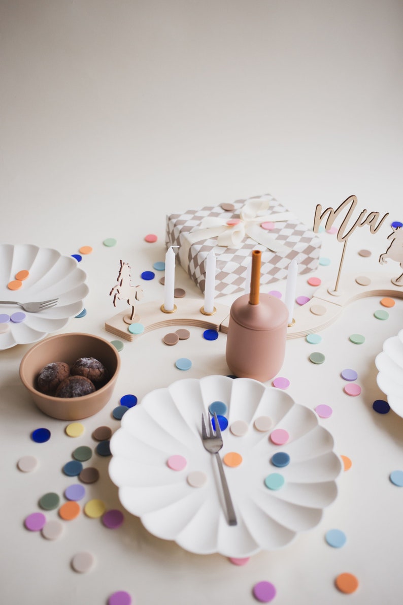 May include: A birthday party table setting with white plates, a brown bowl with chocolate candies, a wooden candle holder with white candles, a gift wrapped in a brown and white checkered paper, a brown and orange silicone cup, and colorful confetti scattered on the table. The name "Mia" is written on a wooden sign.