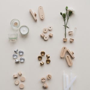 Könnte beinhalten: Eine Auswahl an kleinen Bastelmaterialien aus Holz und Metall, darunter Kerzenhalter, Perlen und dekorative Elemente. Mehrere kleine weiße Kerzen und ein Zweig weißer Blumen sind ebenfalls vorhanden.