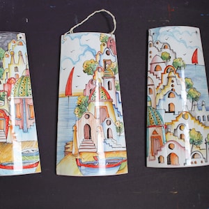 Könnte beinhalten: Drei Keramik-Wandplatten, die farbenfrohe Szenen einer Küstenstadt mit Gebäuden, Bäumen und Booten darstellen. Die Platten sind in einem skurrilen Stil mit leuchtenden Farben und Details bemalt.