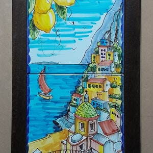 May include: A framed ceramic tile artwork depicting a colorful Italian village with a blue sea and a sailboat in the foreground. The top tile features a lemon tree with yellow lemons and green leaves against a blue sky. The bottom tile features the village with yellow, red, and blue buildings. The text "Positano" is written in blue at the bottom of the artwork.