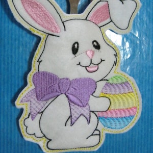 May include: White felt Easter bunny with pink ears, holding a rainbow Easter egg. The bunny is wearing a purple bow and has a yellow outline. The bunny is holding a fork and spoon.