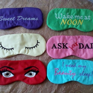 Puede incluir: Cinco antifaces para dormir de colores con frases bordadas. Los antifaces están hechos de tela suave y tienen cintas elásticas. Las frases en los antifaces son "Sweet Dreams", "Wake me at NOON", "ASK your DAD", "I need my Beauty sleep!" y un antifaz con dos ojos grandes.