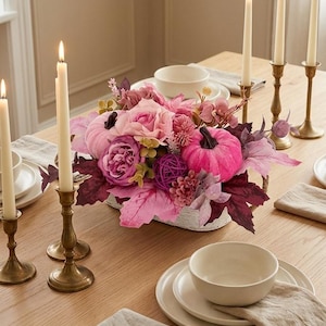 Può includere: Un centrotavola con zucche in velluto rosa, rose artificiali viola e rosa e foglie d'acero bordeaux scuro in un cesto intrecciato bianco, circondato da candelabri in ottone con candele accese su un tavolo da pranzo in legno.