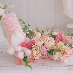 Può includere: Una scatola di legno rosa traboccante di fiori finti nei toni del rosa e del crema, accentuata da foglie verdi. Uno gnomo rosa con un cappello di lana e una barba bianca è seduto accanto a un cuore rosa intrecciato. La scatola ha del testo.