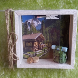 May include: A white wooden shadow box with a picture of a small cabin in the mountains. The picture is surrounded by a brown rope and a small green backpack is in the foreground.