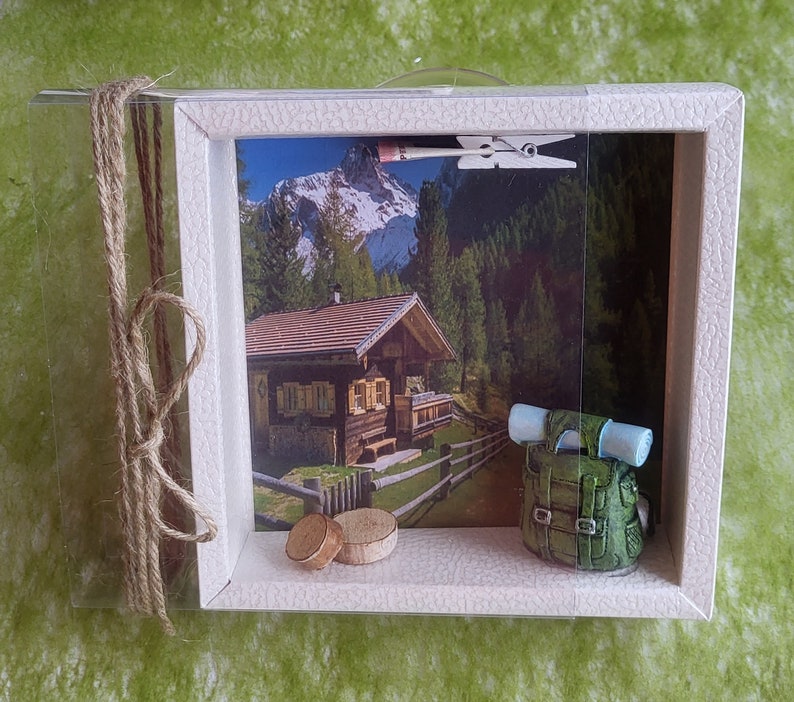 May include: A white wooden shadow box with a picture of a small cabin in the mountains. The picture is surrounded by a brown rope and a small green backpack is in the foreground.