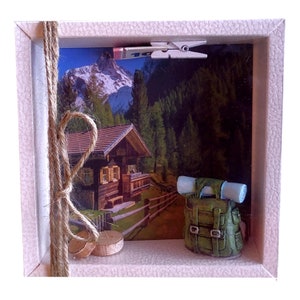 May include: A small wooden frame with a picture of a mountain cabin and a green backpack with a blue sleeping bag. There are two wooden circles and a piece of twine in the frame.