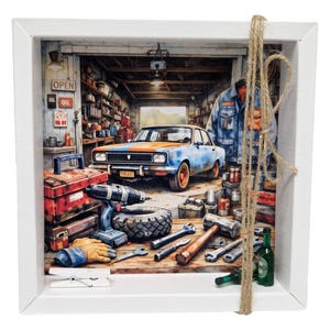 Könnte beinhalten: Ein gerahmtes Kunstwerk, das eine Garagenszene mit einem blau-orangen Auto zeigt. Werkzeuge, Reifen und eine Werkzeugkiste befinden sich im Vordergrund. Das Wort "OIL" ist auf einem Schild zu sehen. Das Kunstwerk ist in einem weißen quadratischen Rahmen mit einem Bindfadenakzent gerahmt.