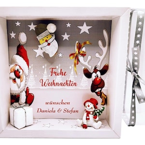 Könnte beinhalten: Weiß gerahmte Weihnachtsschachtel mit Santa Claus, Rentier und Schneemann. Der Text "Frohe Weihnachten" und Namen sind abgebildet. Der Hintergrund ist grau mit weißen Sternen und Weihnachtsbäumen.