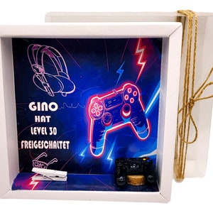 Könnte beinhalten: Eine weiße Schattenbox mit blauem und schwarzem Hintergrund, auf dem sich ein neonpinkfarbener Videospiel-Controller und ein Kopfhörer befinden. Der Text "Gino Hat Level 30 Freigeschaltet" ist auf den Hintergrund gedruckt.