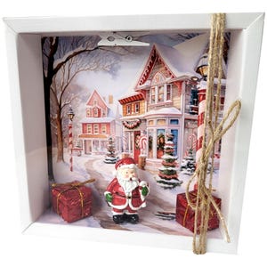 Könnte beinhalten: Eine weihnachtliche Schattenbox mit einer Miniatur-Weihnachtsmannfigur in rotem Anzug, vor einer verschneiten Dorflandschaft. Zwei rote Geschenkboxen und eine Schnurschleife ergänzen die festliche Szene. Die Box ist weiß.
