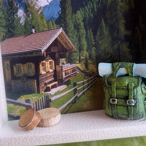 May include: A miniature wooden cabin with a red roof and a green backpack with a blue roll on top, sitting on a shelf with two wooden discs.