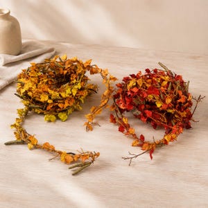 Könnte beinhalten: Zwei dekorative Girlanden aus ineinander verschlungenen braunen Zweigen und künstlichen Herbstblättern. Eine Girlande zeigt gelbes und oranges Laub, während die andere leuchtend rote, orange und gelbe Blätter aufweist. Beide enthalten kleine grüne Kätzchen.
