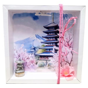 Könnte beinhalten: Weißer Schattenkasten mit einer japanischen Pagoden-Szene, dem Berg Fuji im Hintergrund und rosa Kirschblüten. Eine kleine Steinlaterne befindet sich im Vordergrund. Eine rosa Schnurschleife und eine Blume schmücken die Box.