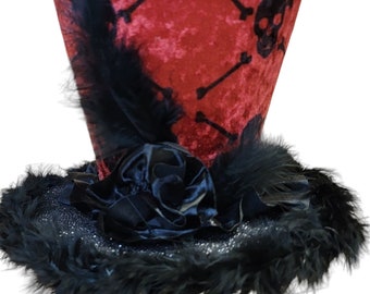 Peut inclure: Un chapeau haut de forme en velours rouge avec un motif de crâne et d'os croisés noirs, des plumes noires et une rose en velours noir.