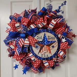 Op de afbeelding: Een rood, wit en blauwe patriottische krans met een stervormig bord waarop "God Bless the U.S.A." staat.