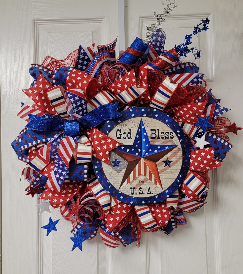 Op de afbeelding: Een rood, wit en blauwe patriottische krans met een stervormig bord waarop "God Bless the U.S.A." staat.