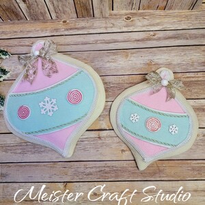 May include: Two felt Christmas ornaments in the shape of teardrops. The larger ornament is pink with a light blue center and the smaller ornament is pink with a light blue center. Both ornaments have white snowflakes, candy cane swirls, and gold glitter accents.