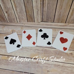 May include: Four white card-shaped decorations with red and black glitter accents. The cards feature the four suits of a standard deck of cards: hearts, diamonds, clubs, and spades. The cards are attached to white sticks.