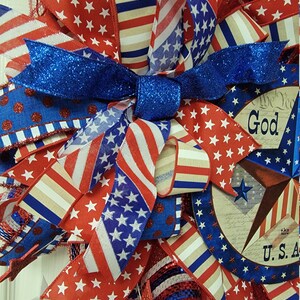 Op de afbeelding: Een patriottische krans met rood, wit en blauw lint, sterren en een bord met de tekst "God Bless the U.S.A."