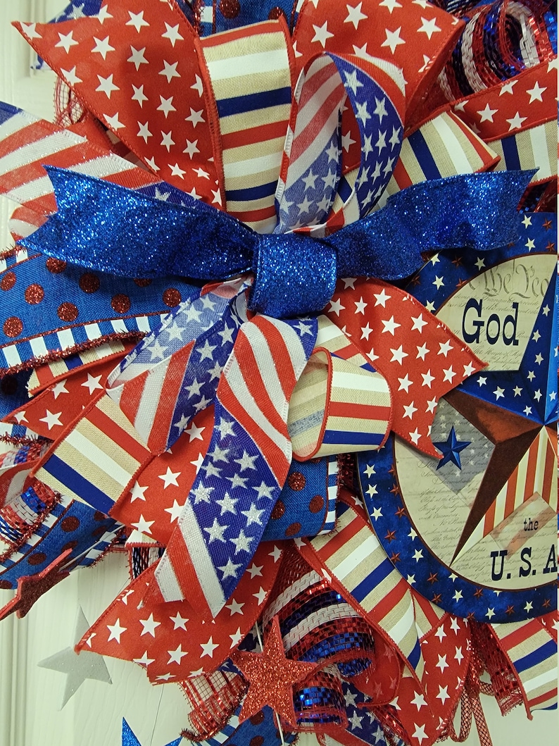 Op de afbeelding: Een patriottische krans met rood, wit en blauw lint, sterren en een bord met de tekst "God Bless the U.S.A."