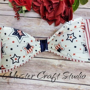 May include: A white bow with red and blue stars and stripes. The bow is tied with a blue ribbon. The bow is on a wooden background. Meister Craft Studio is written on the background.