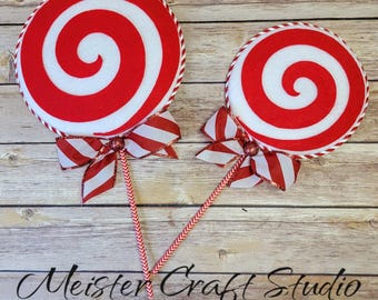 May include: Two decorative red and white swirl lollipops with red and white striped bows and stems. The lollipops have a spiral design and are trimmed with a white and red rope-like border. The text "Meister Craft Studio" is visible on the wooden background.
