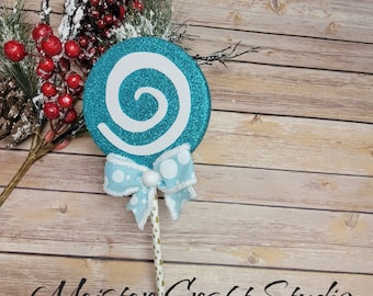 May include: A teal blue glitter swirl lollipop with a white swirl design and a white and blue polka dot bow. The lollipop is on a stick with a gold and white striped pattern.