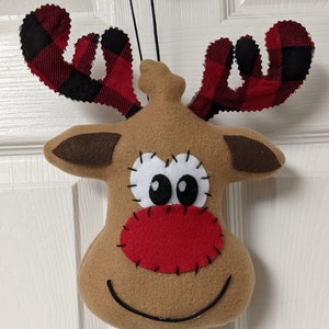 May include: A brown felt reindeer head with red and black plaid antlers. The reindeer has a red felt nose, white felt eyes, and a black stitched smile.
