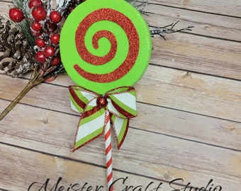 May include: A green and red glitter lollipop with a white and red striped stick. The lollipop is tied with a green and white striped ribbon with a red bow.