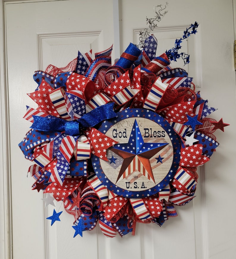 Op de afbeelding: Een rood, wit en blauwe patriottische krans met een stervormig bord met de tekst "God Bless U.S.A."